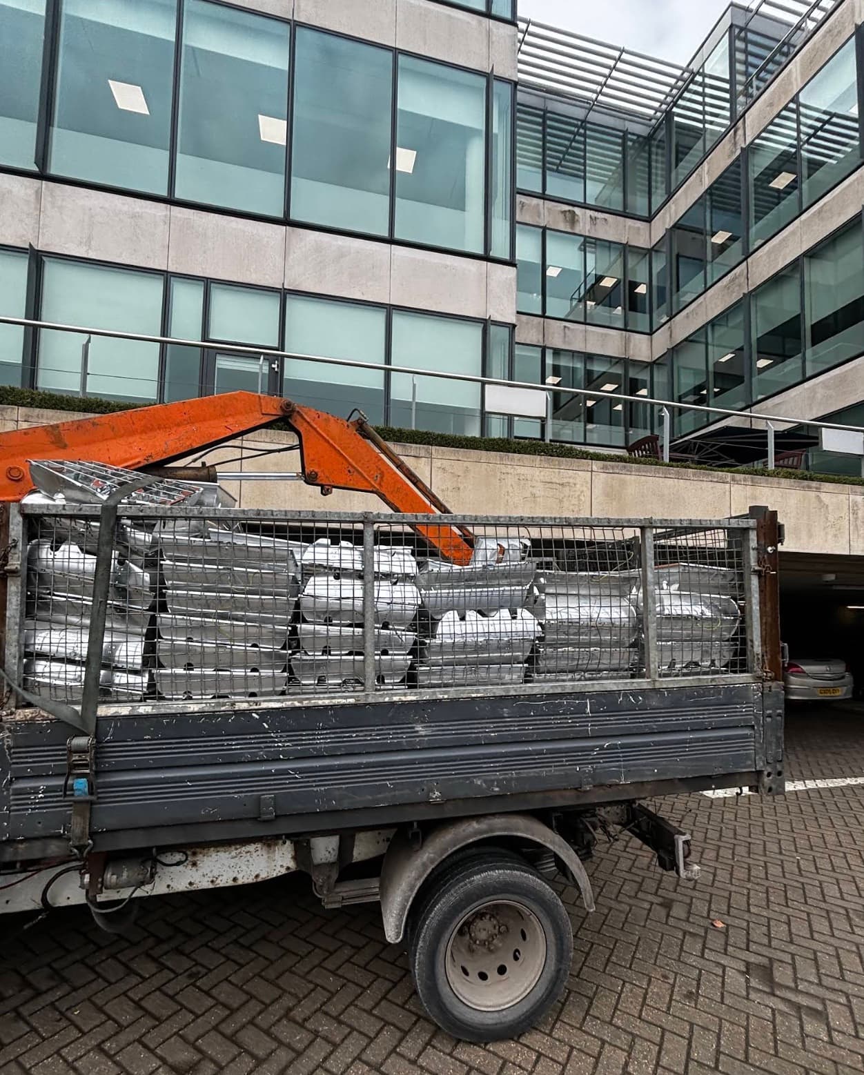 A caged tipper loaded with stripped metal and steel kegs parked outside a commercial office building, HIAB arm raised